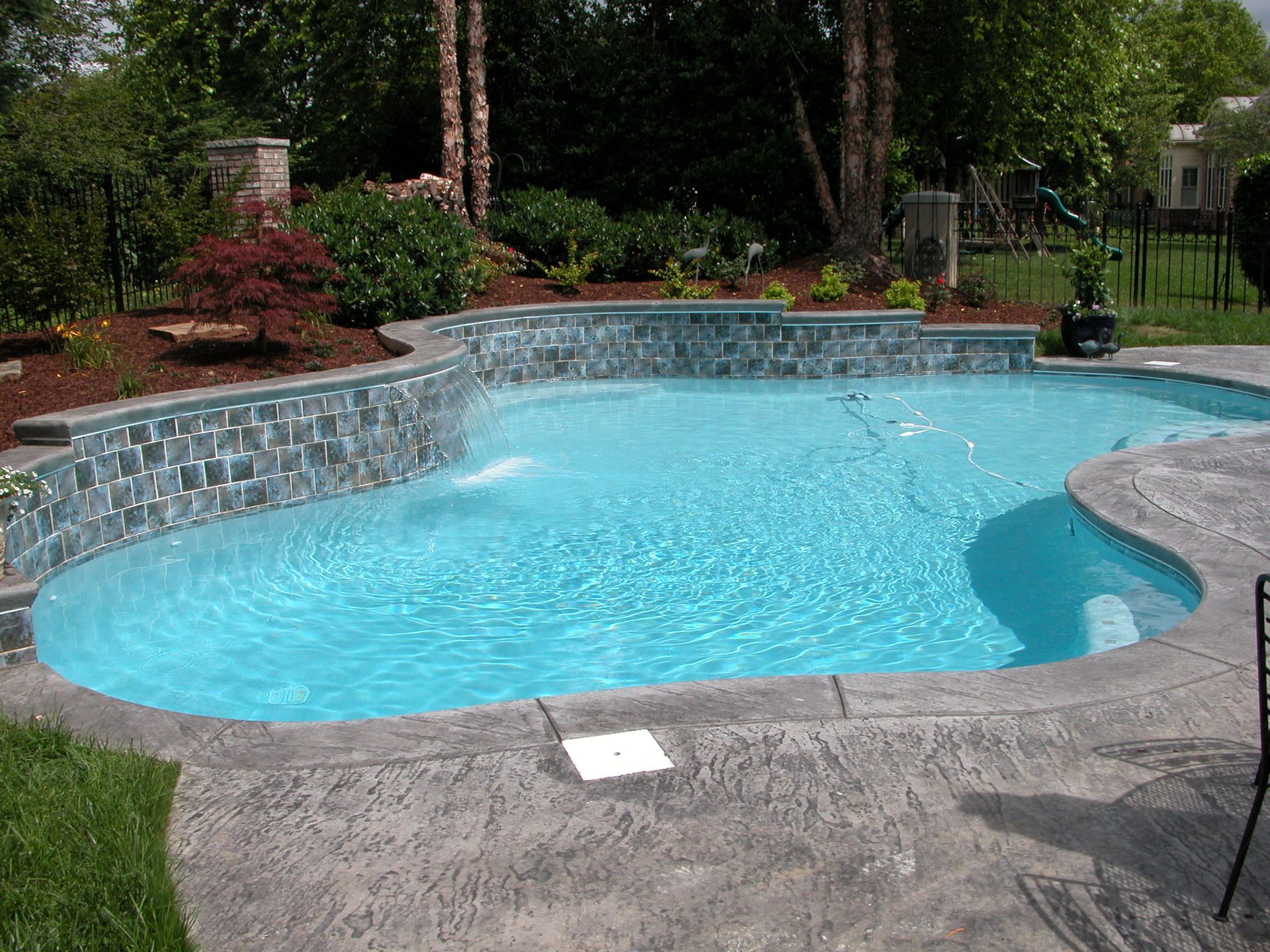 Backyard swimming pool with blue water, stone deck, and landscaped garden surroundings