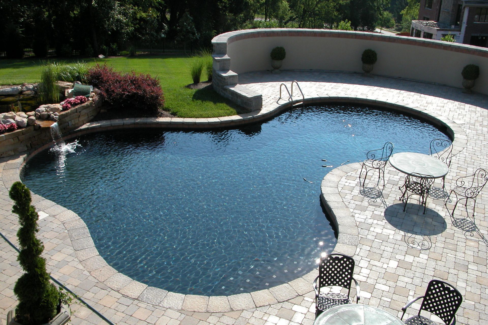 Backyard swimming pool with blue water, stone deck, and patio tables in bright sunlight