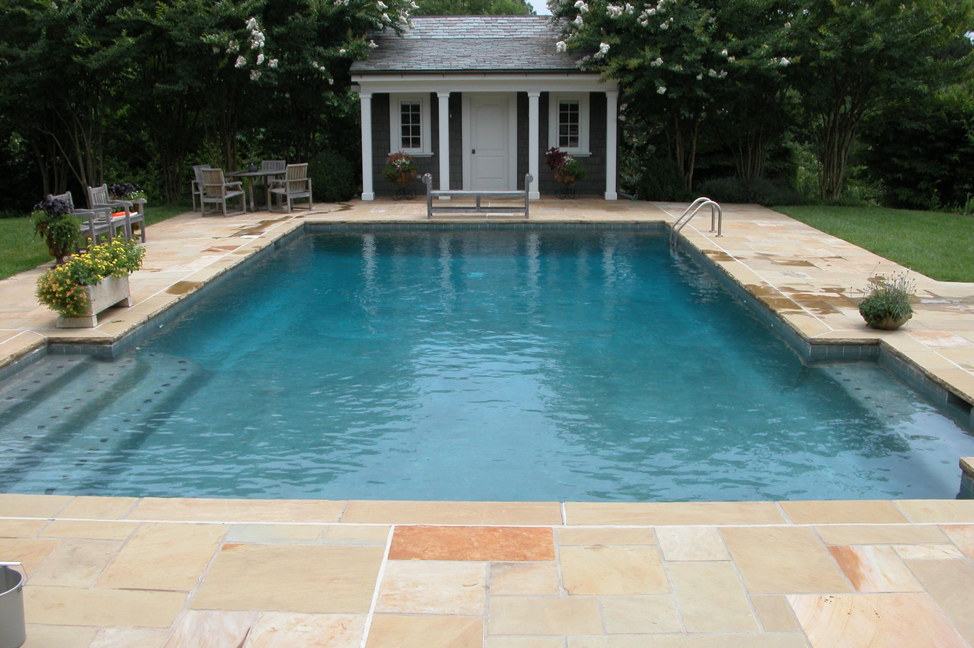 Backyard swimming pool with stone patio and a small white pool house surrounded by trees