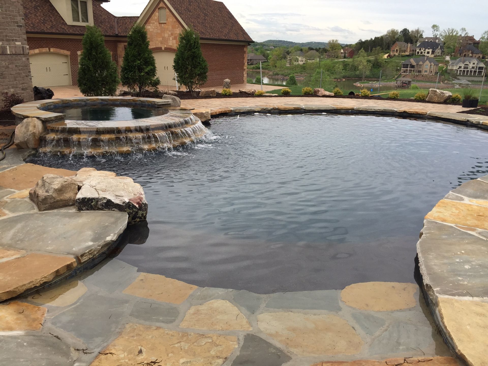 Stone-edged backyard pool with waterfall, calm water, and rustic buildings in the background