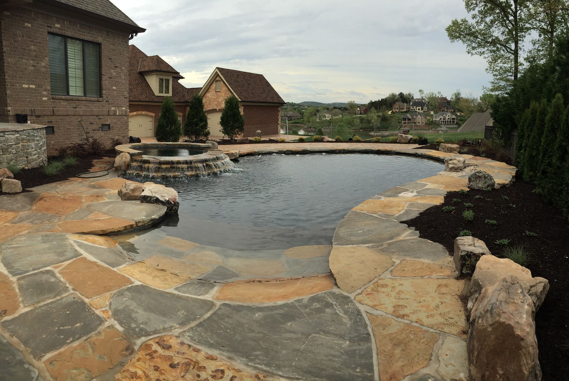 Stone backyard pool with flagstone deck beside brick homes and trees