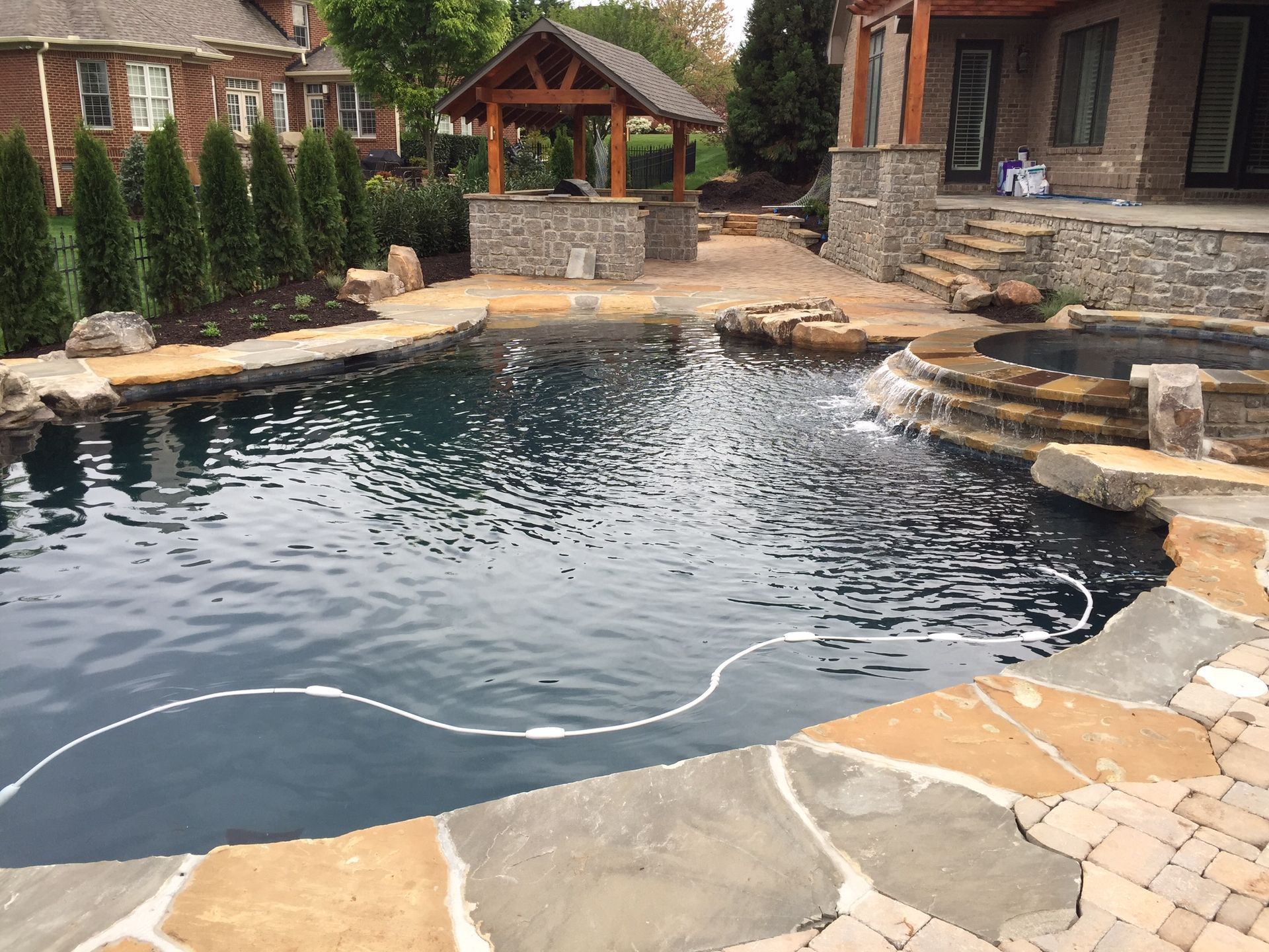 Backyard stone pool with dark water, rock edge, and a small pavilion beside a brick house.