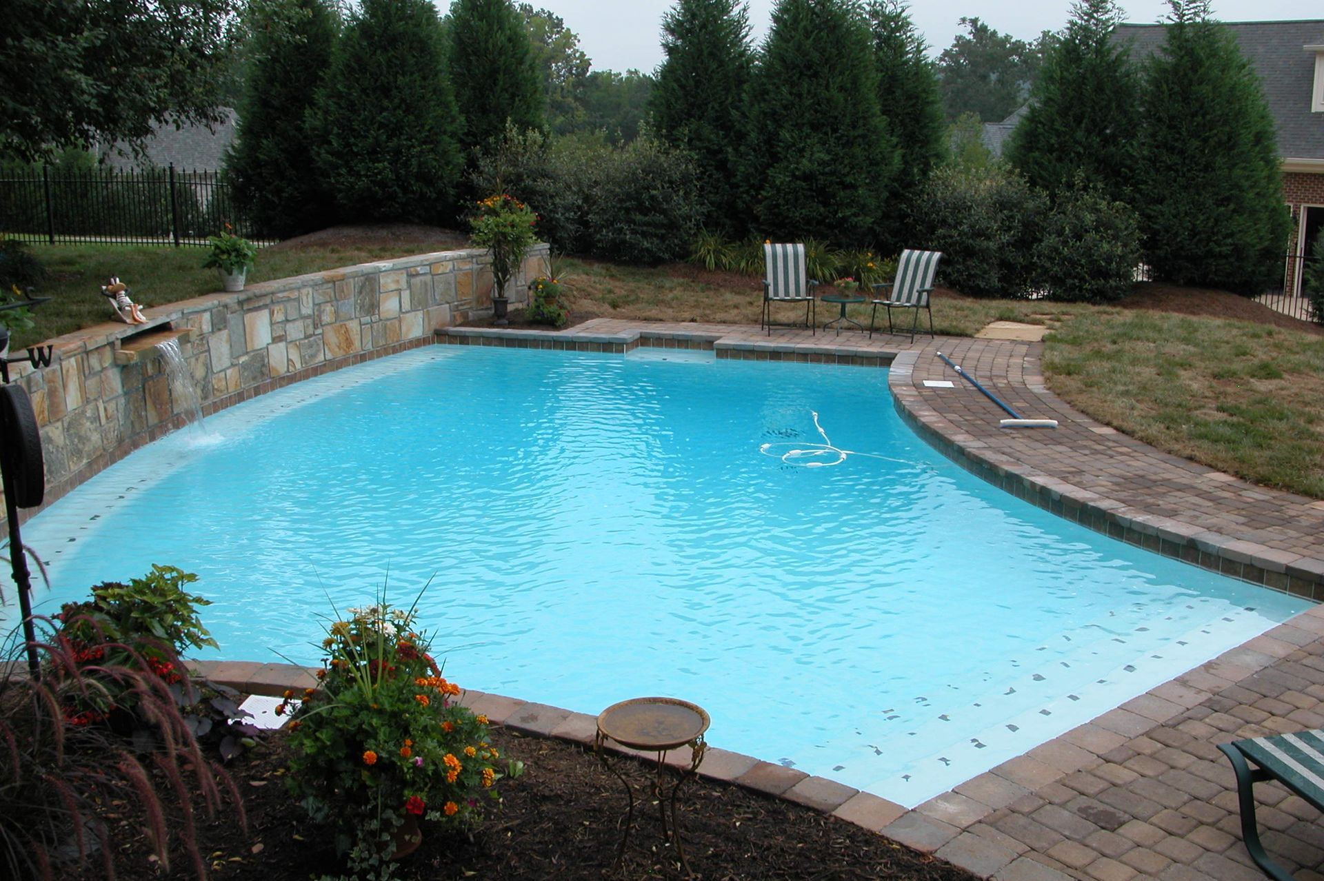Backyard swimming pool with clear blue water, stone patio, and surrounding trees and shrubs