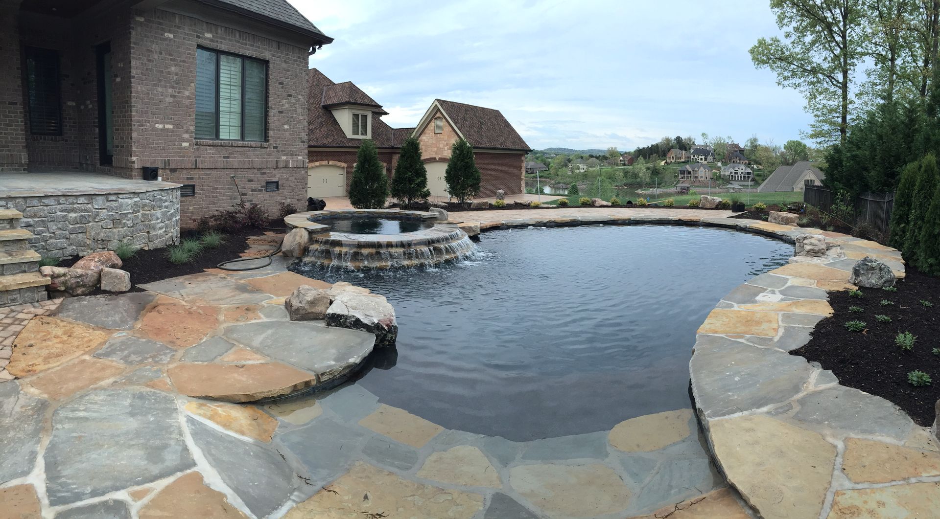 Stone pool and patio beside a brick house, with a calm dark water feature and landscaped rocks.
