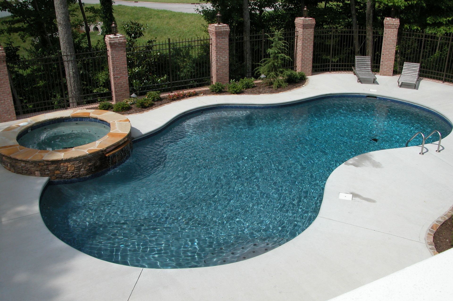 Backyard swimming pool with attached hot tub and stone deck, bordered by brick columns and greenery