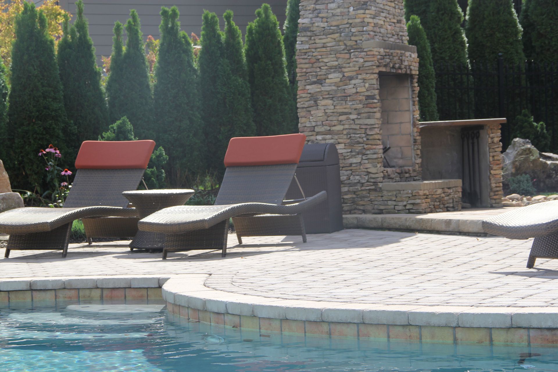 Poolside lounge chairs beside a stone patio and fireplace, with tall hedges in the background