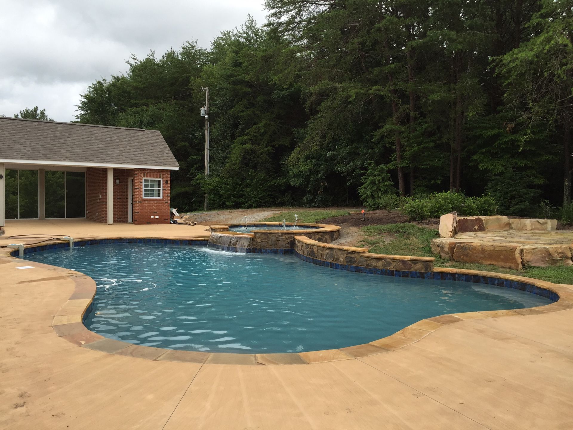 Backyard swimming pool with tan deck, stone waterfall, and a small pool house beside trees