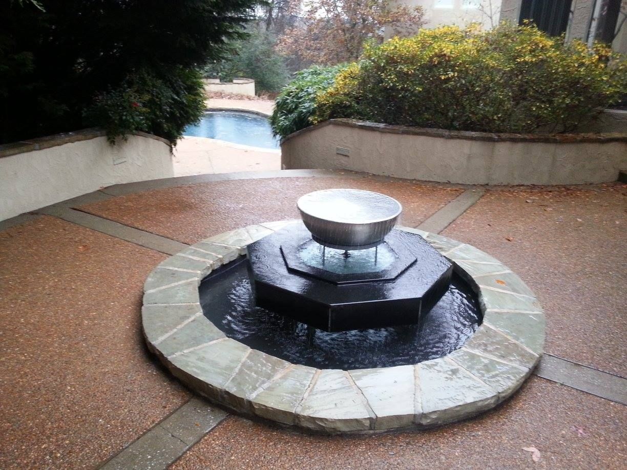 Circular stone fountain in a landscaped courtyard with a small metal bowl in the center.