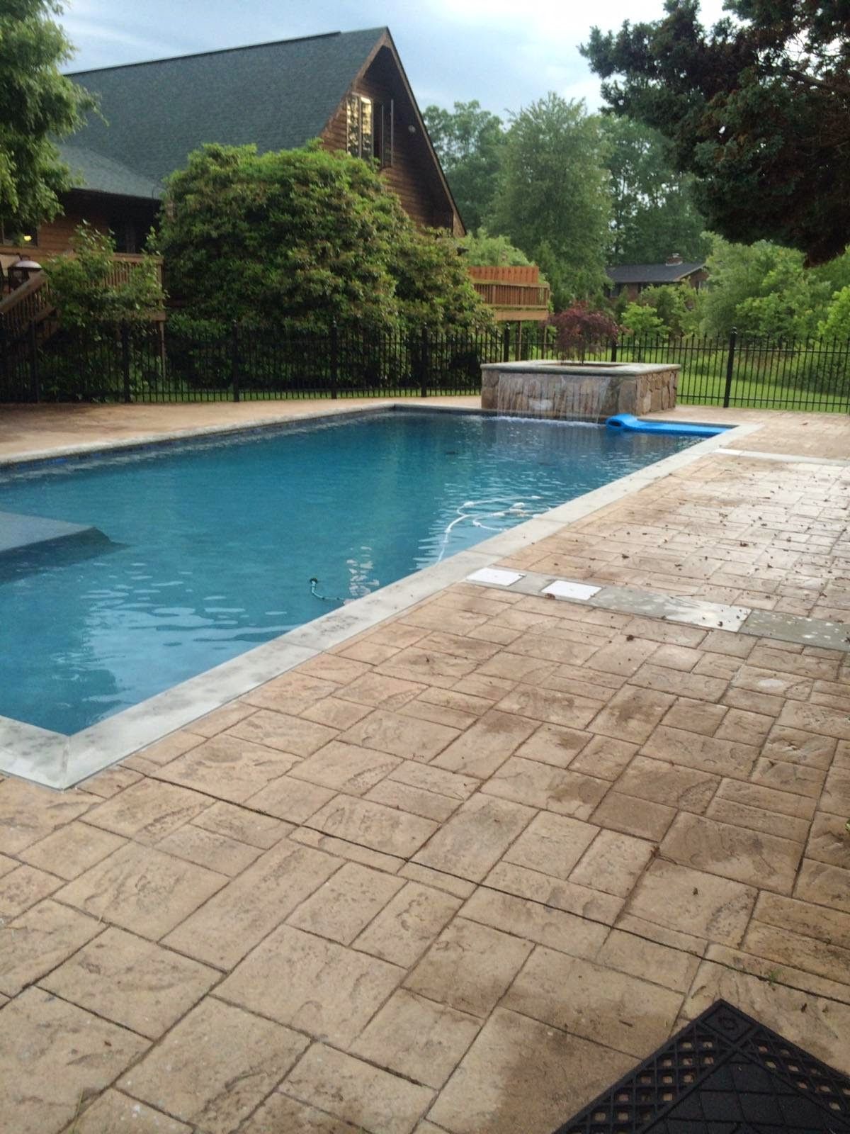 Backyard swimming pool beside a stone patio and house, with greenery and trees nearby