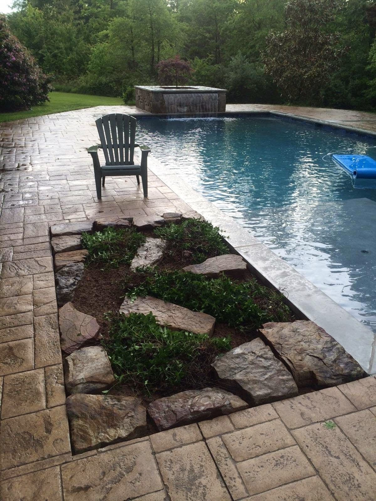 Stone patio with a chair beside a curved blue swimming pool and lush green garden.