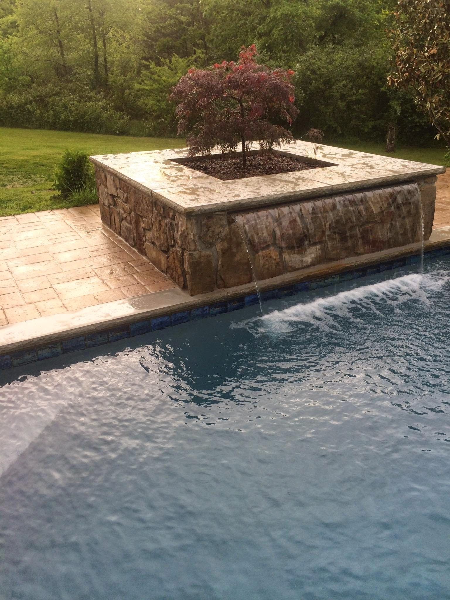 Stone poolside planter with a small red shrub beside blue water and green landscaping