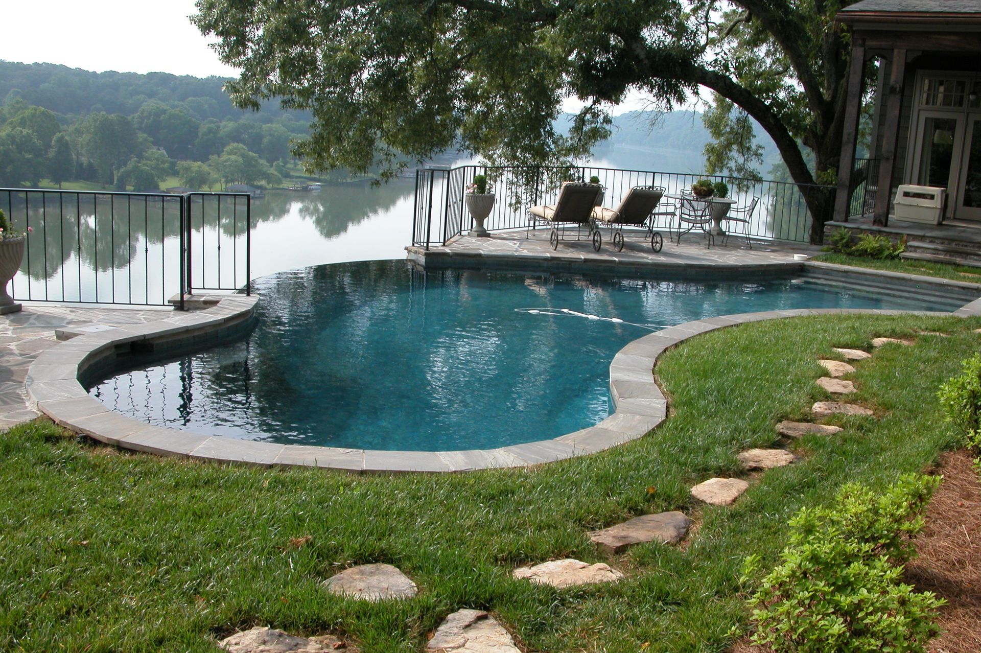 Curved backyard pool beside a lake, with lounge chairs, grass, and trees in the background