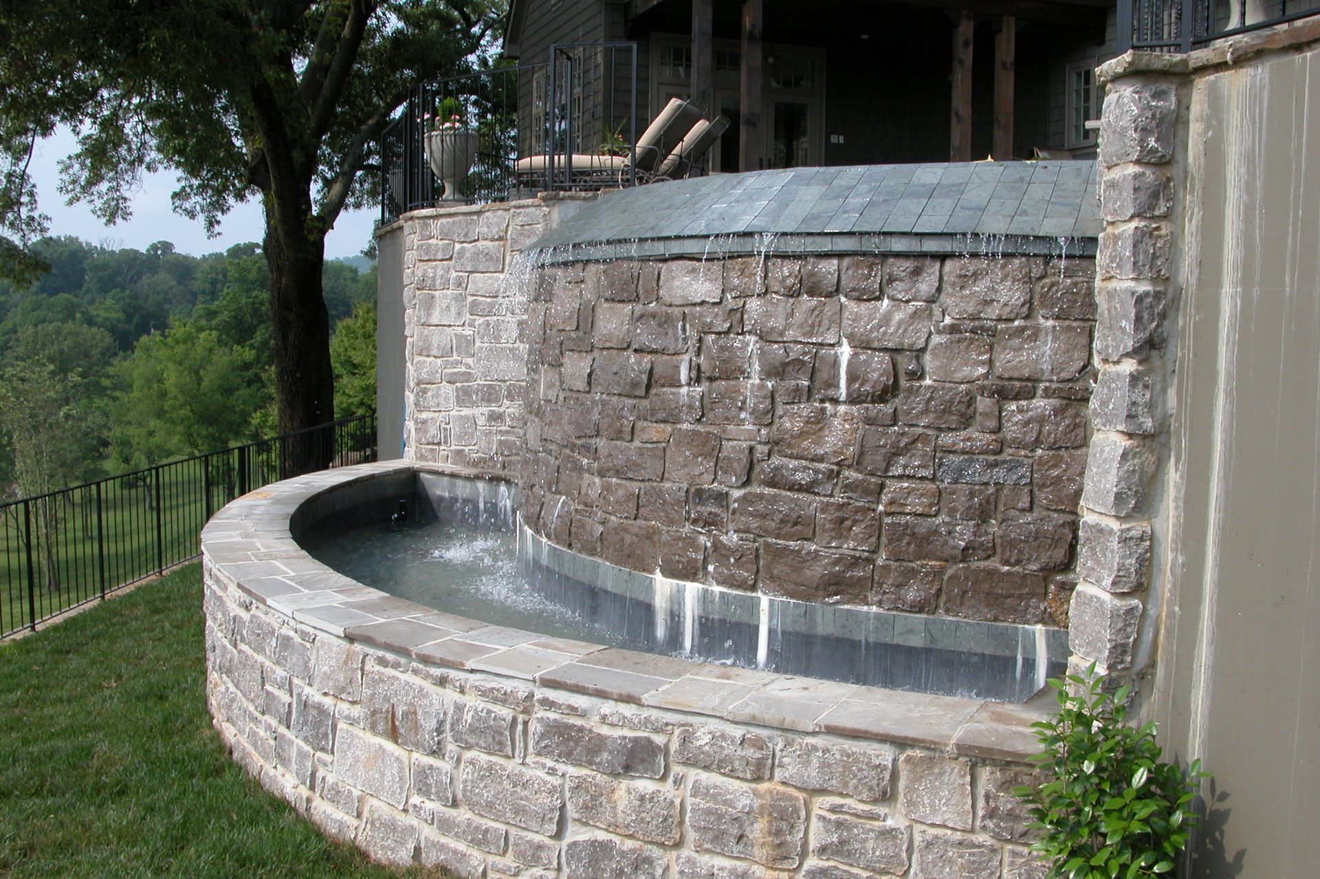 Curved stone fountain wall with flowing water beside a wooded hillside and patio