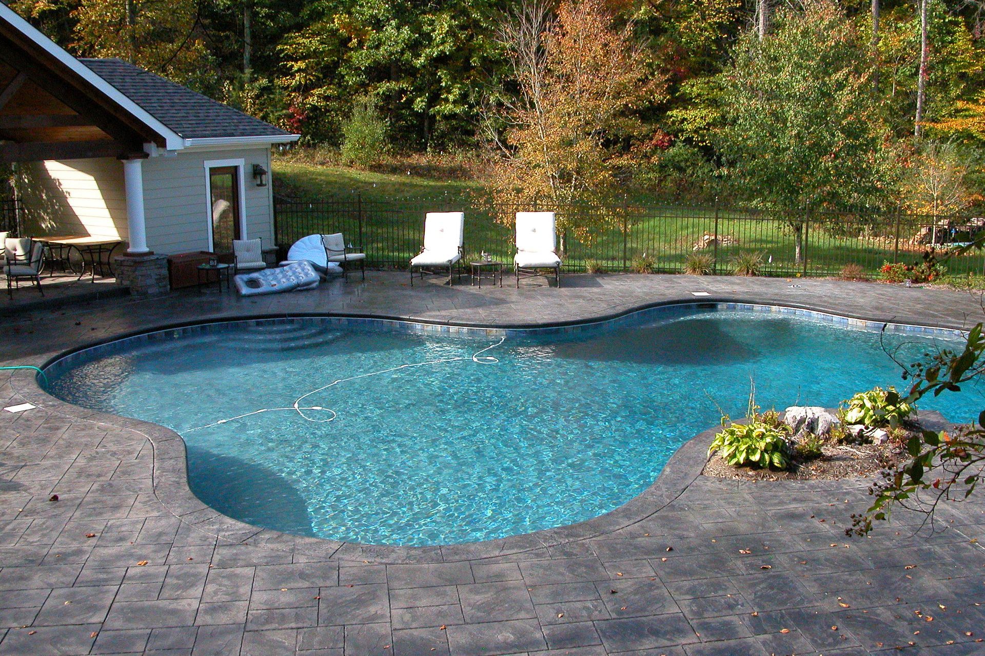 Backyard swimming pool with stone deck, lounge chairs, and flower bed in a wooded setting
