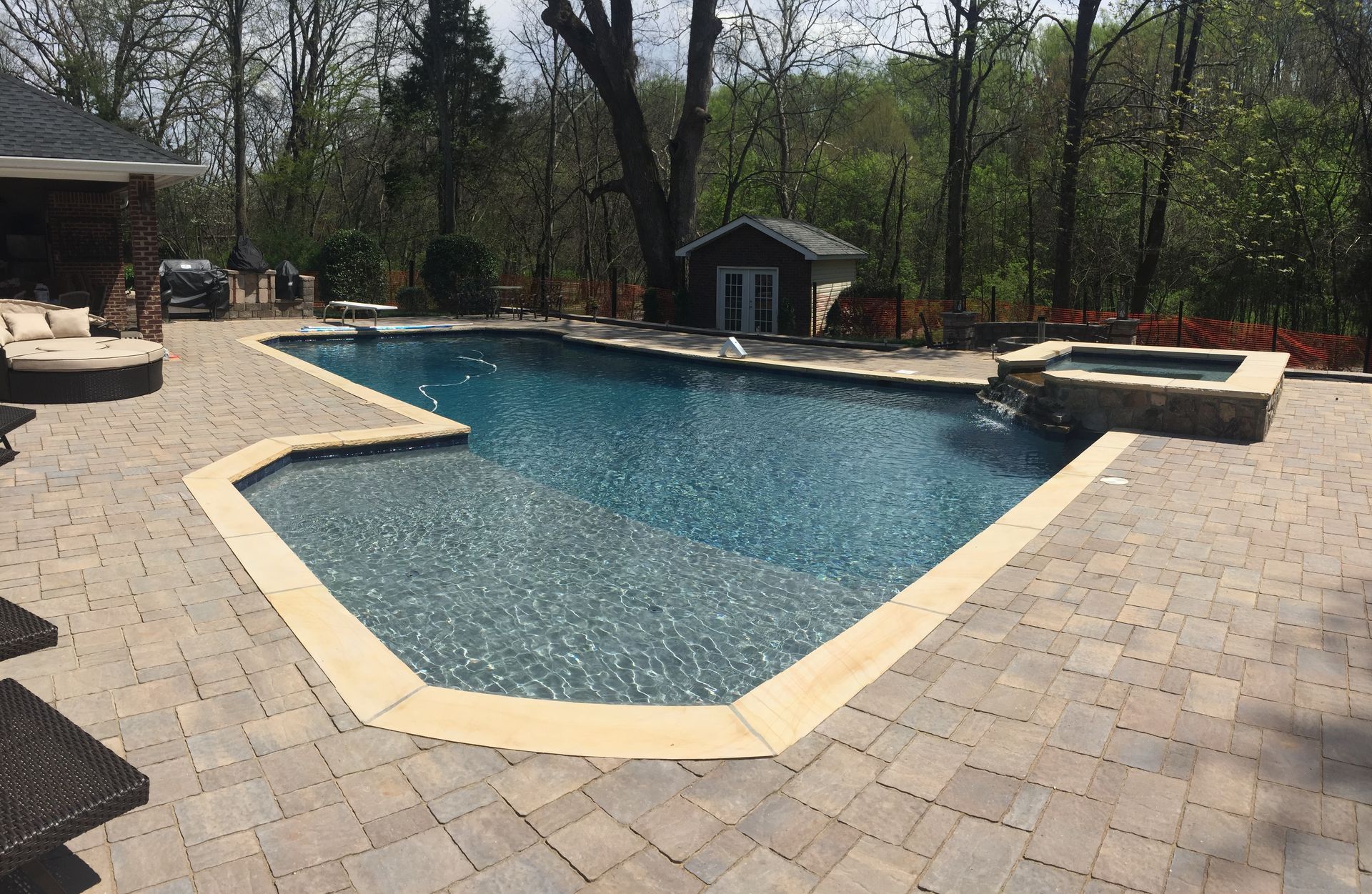 Backyard swimming pool with tan paver deck, lounge chairs, and wooded trees in the background