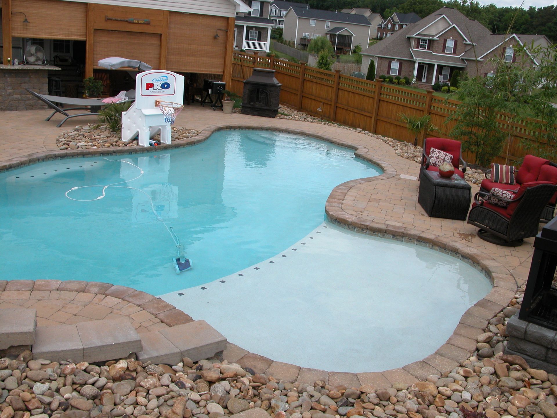 Backyard pool with shallow area, patio chairs, and a robotic pool cleaner near the water's edge