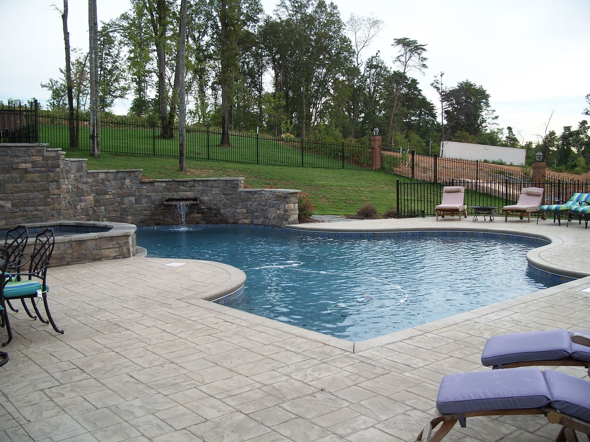 Backyard pool with stone patio, water feature, lounge chairs, and a grassy fenced yard