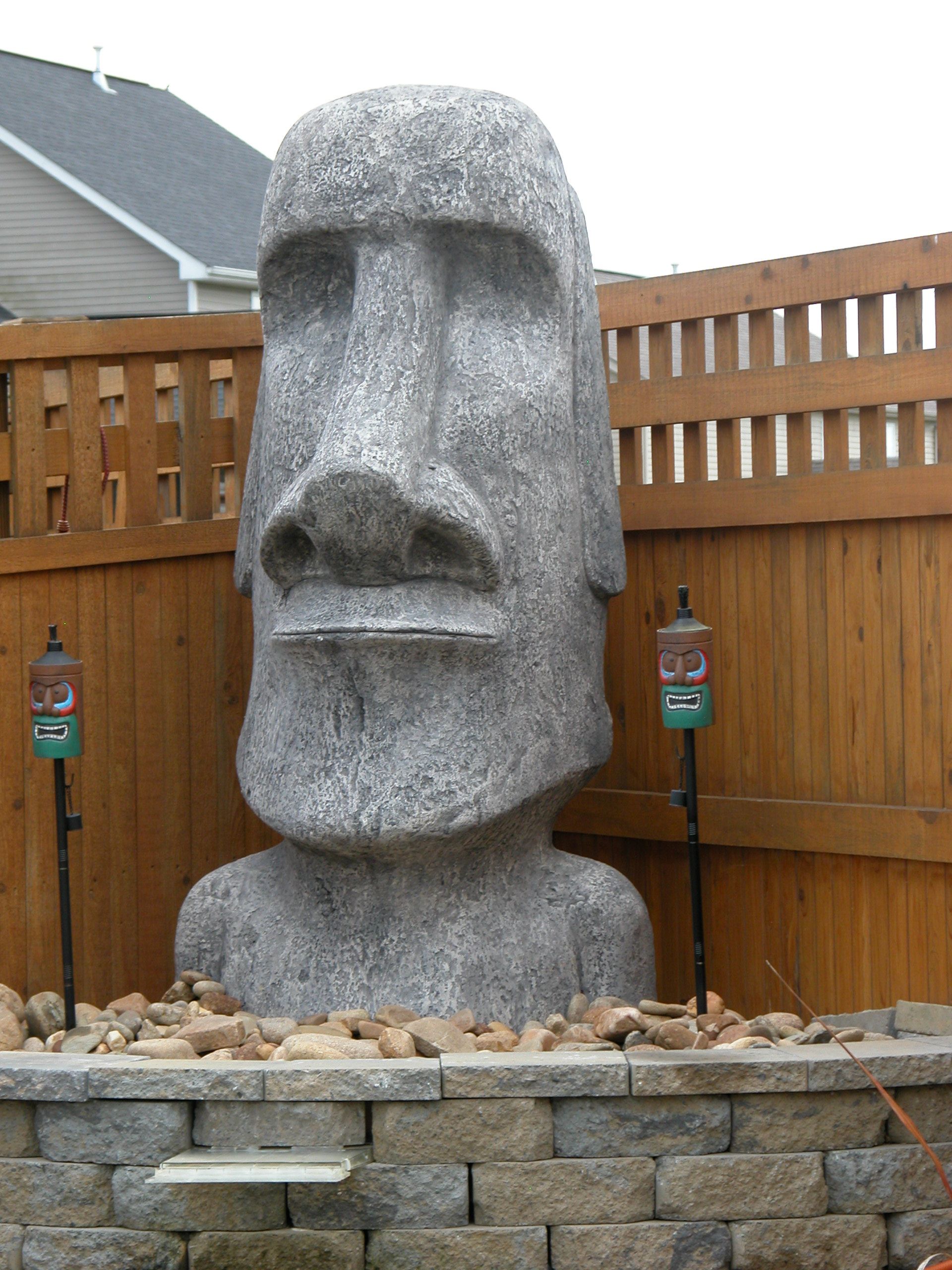 Large gray stone Moai-style statue in a fenced yard behind a low stone wall.
