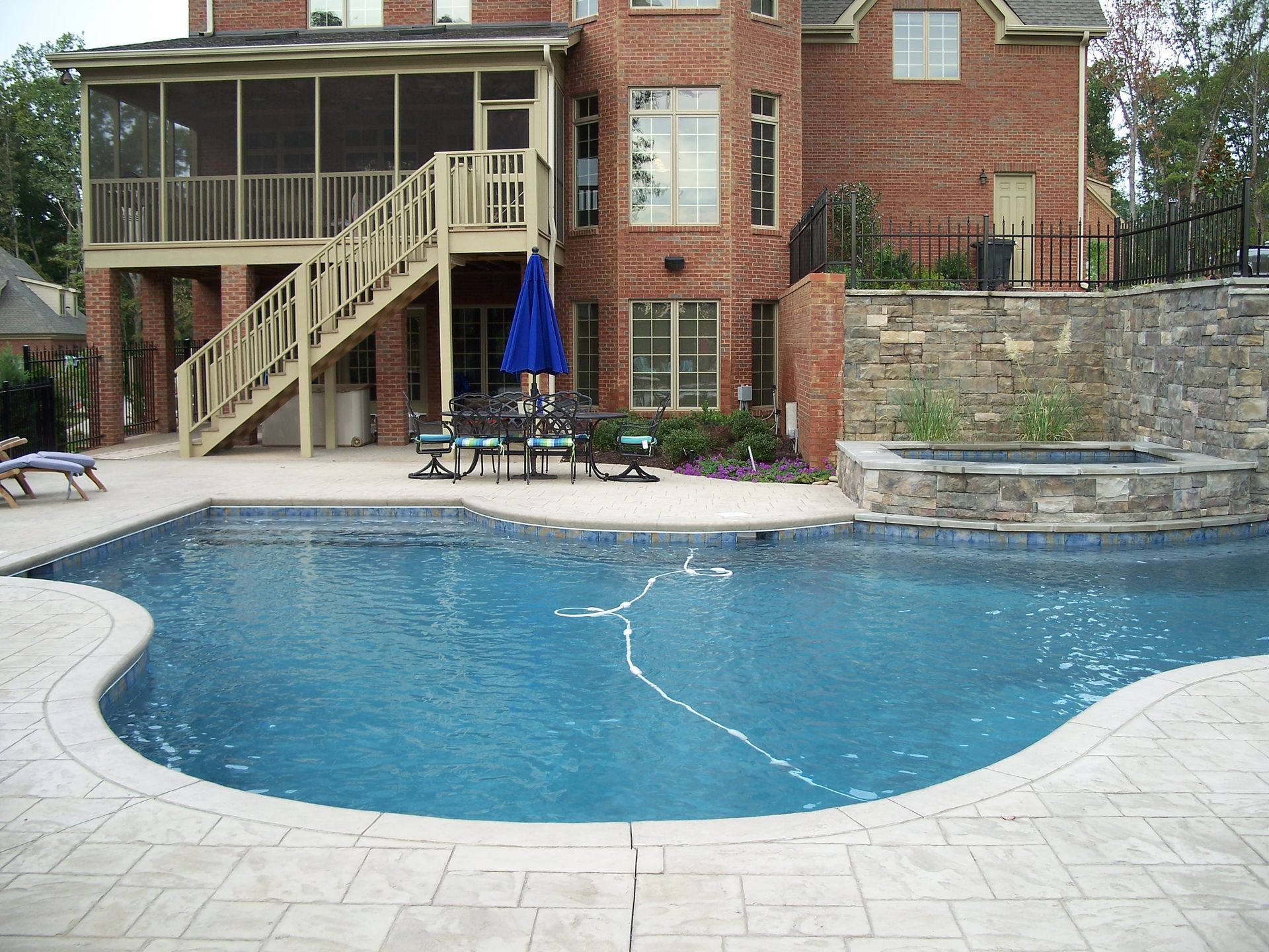 Backyard swimming pool beside a brick house with patio stairs, umbrella, and stone retaining wall