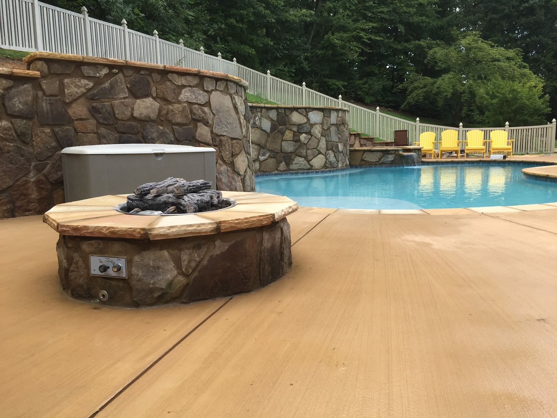 Backyard pool deck with a stone fire pit and yellow lounge chairs beside the water