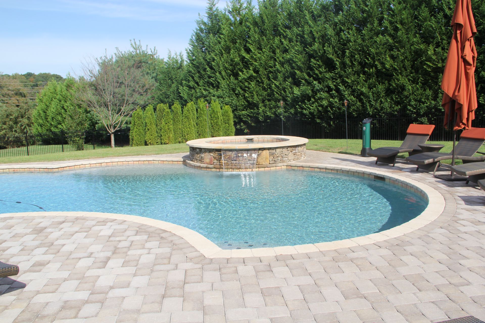 Backyard pool with stone deck, spa, lounge chairs, and orange umbrella under a sunny sky