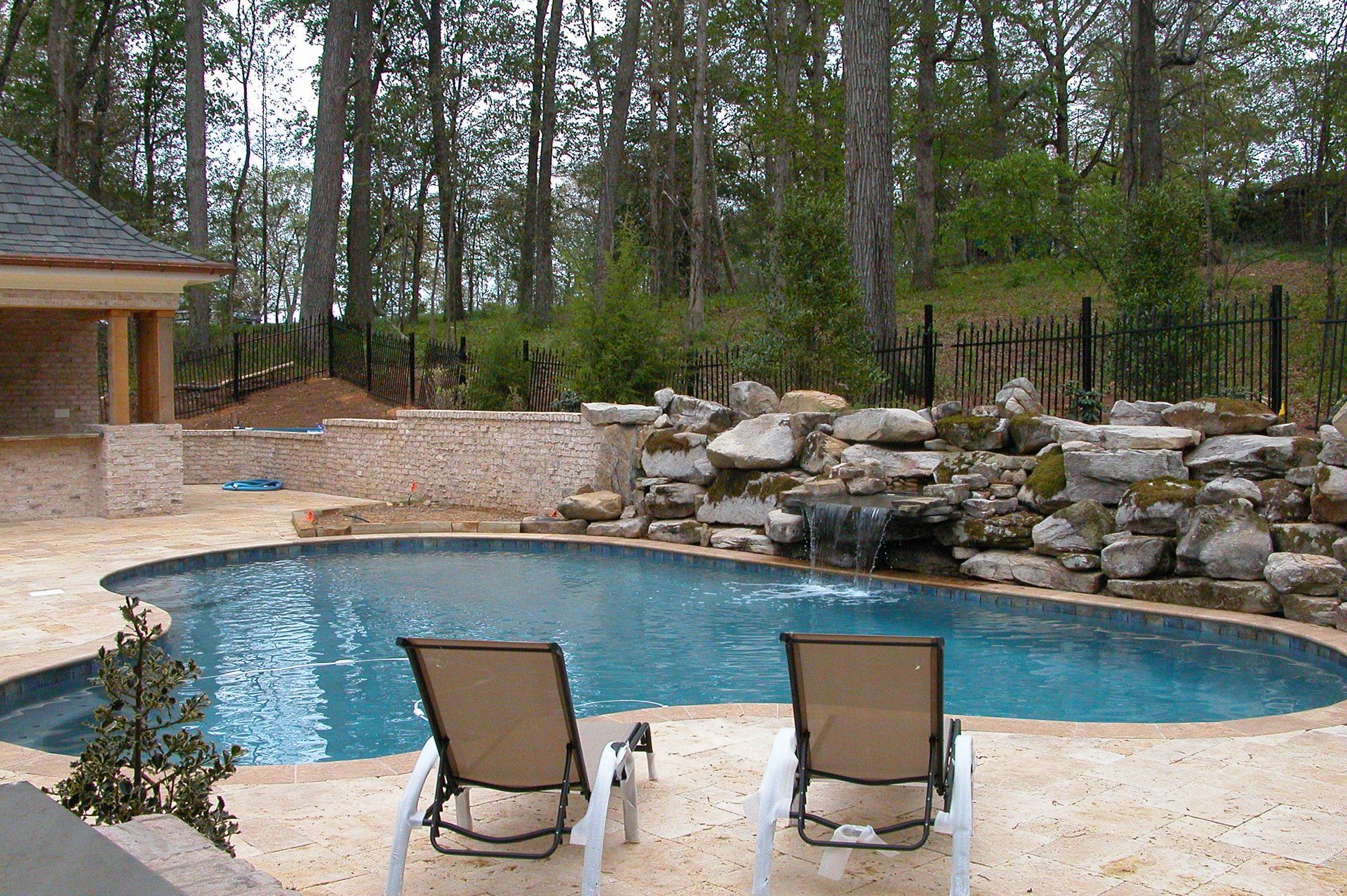 Backyard pool with two lounge chairs, stone waterfall, and wooded landscaping