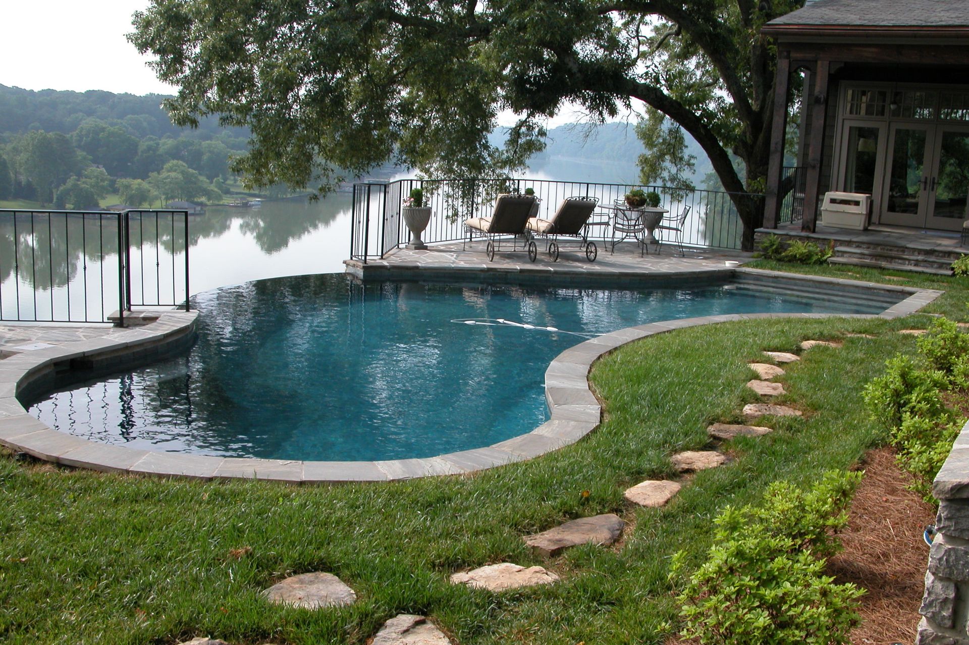 Curved backyard pool with stone deck, green lawn, and lake view at sunset