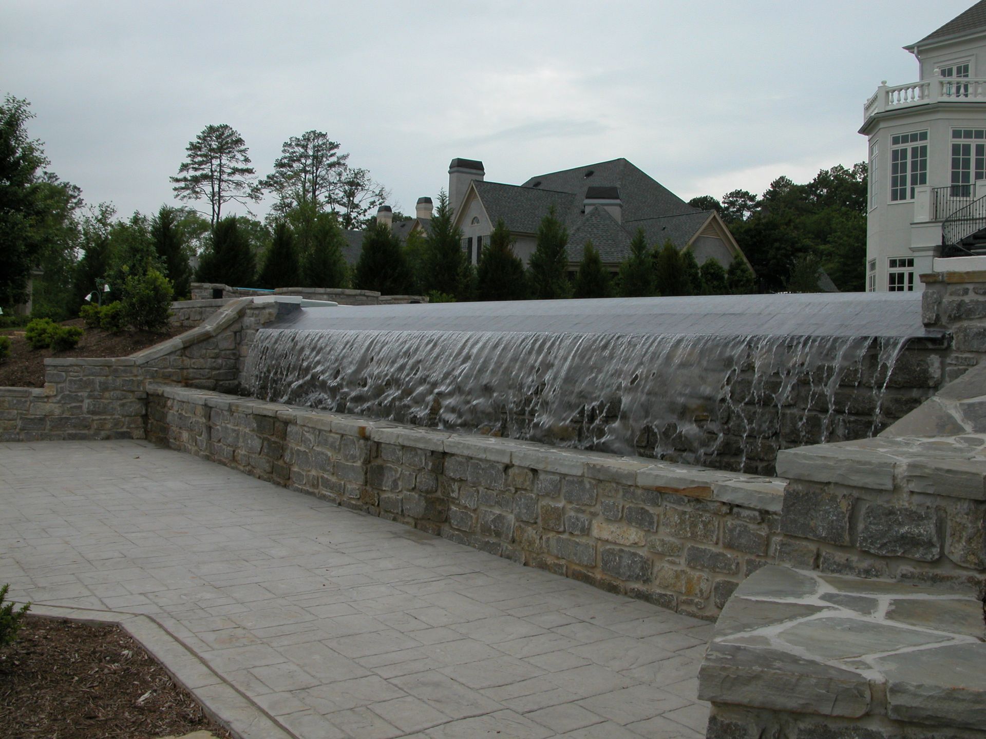 Stone waterfall wall along a paved path, with trees and buildings in the background under a cloudy sky