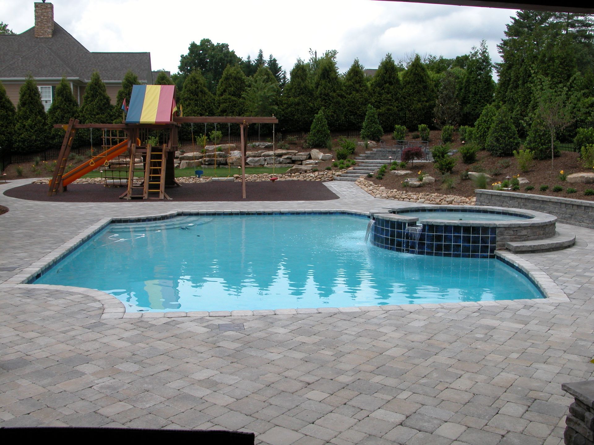 Backyard pool with stone patio, curved hot tub, and a colorful playset in the background