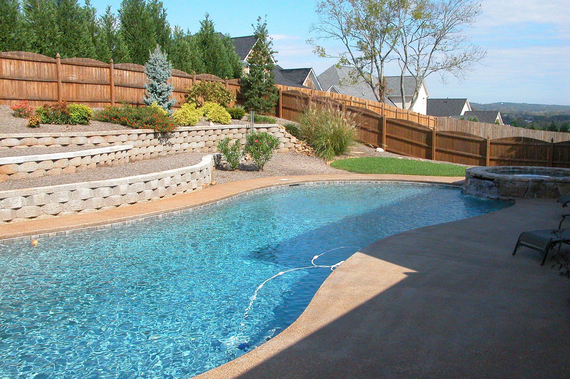 Backyard swimming pool with blue water, stone landscaping, wooden fence, and patio seating under a clear sky
