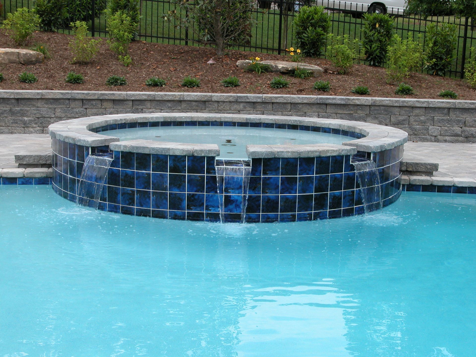 Circular tiled hot tub beside a blue swimming pool with landscaped greenery behind it