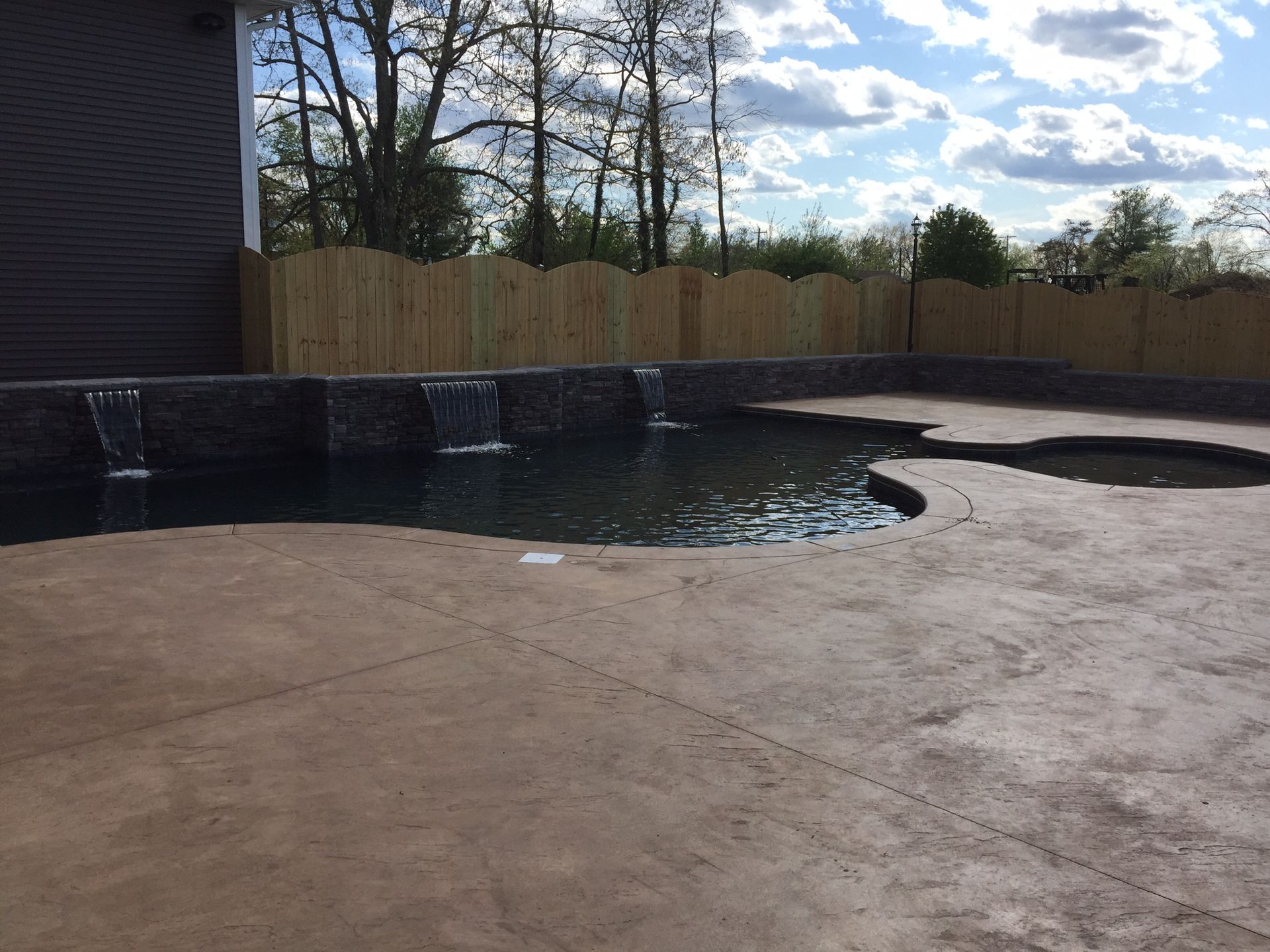 Empty backyard pool with dark water, concrete deck, wooden fence, and cloudy sky.