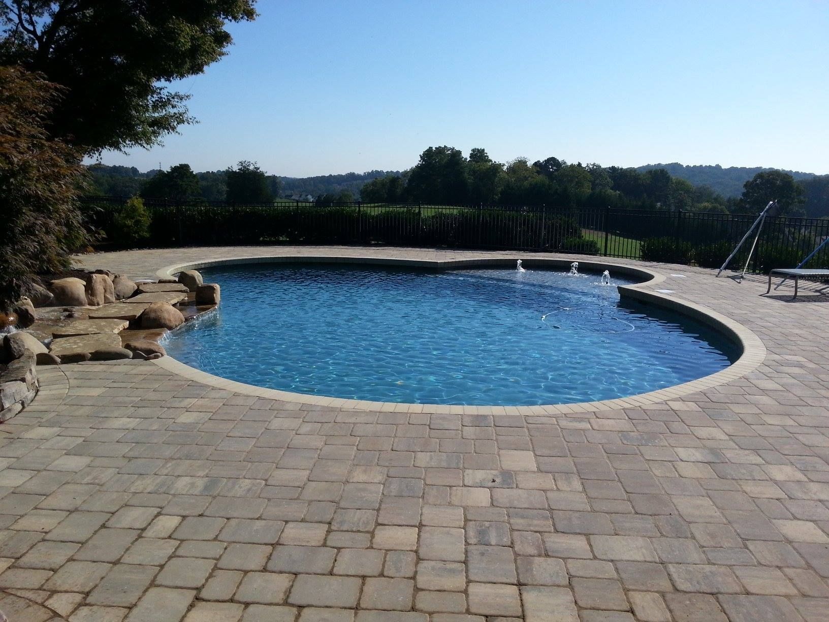 Curved backyard swimming pool with blue water, stone patio, and distant trees under a clear sky