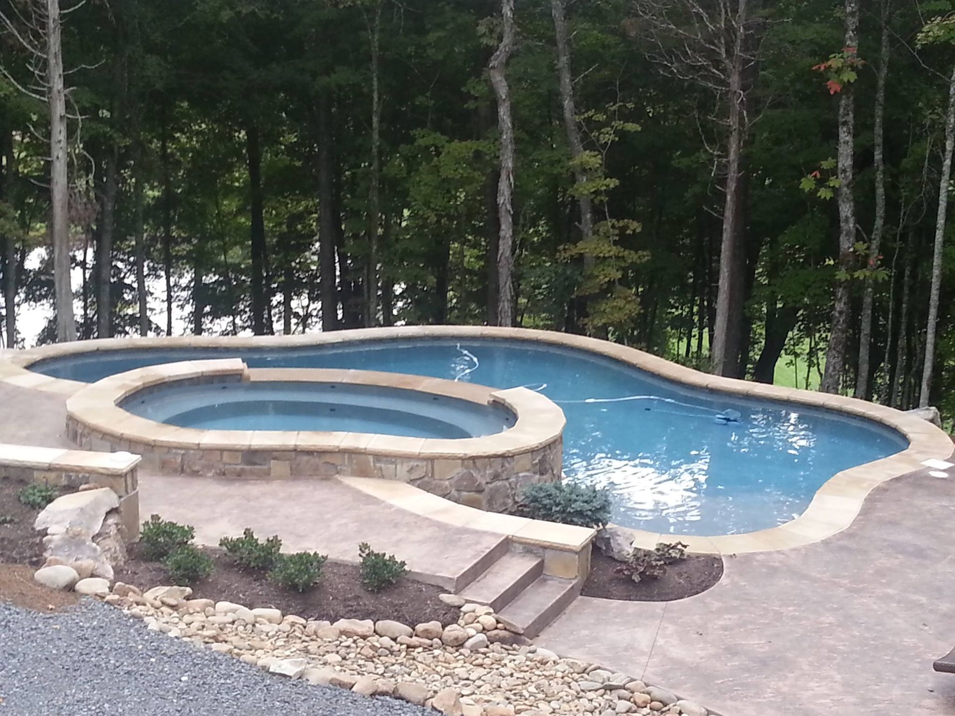 Backyard pool with attached hot tub, stone patio, and trees in the background