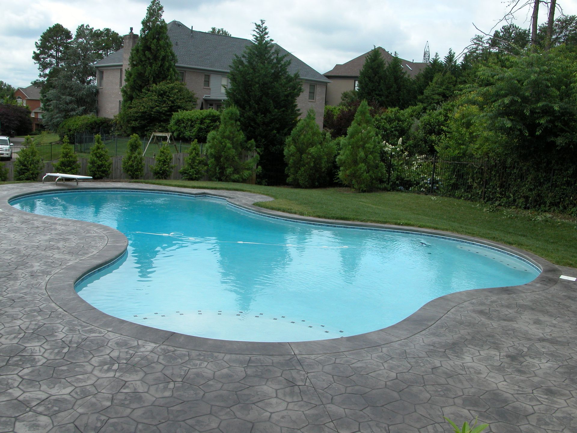 Backyard swimming pool with curved stone deck and lush green trees around it