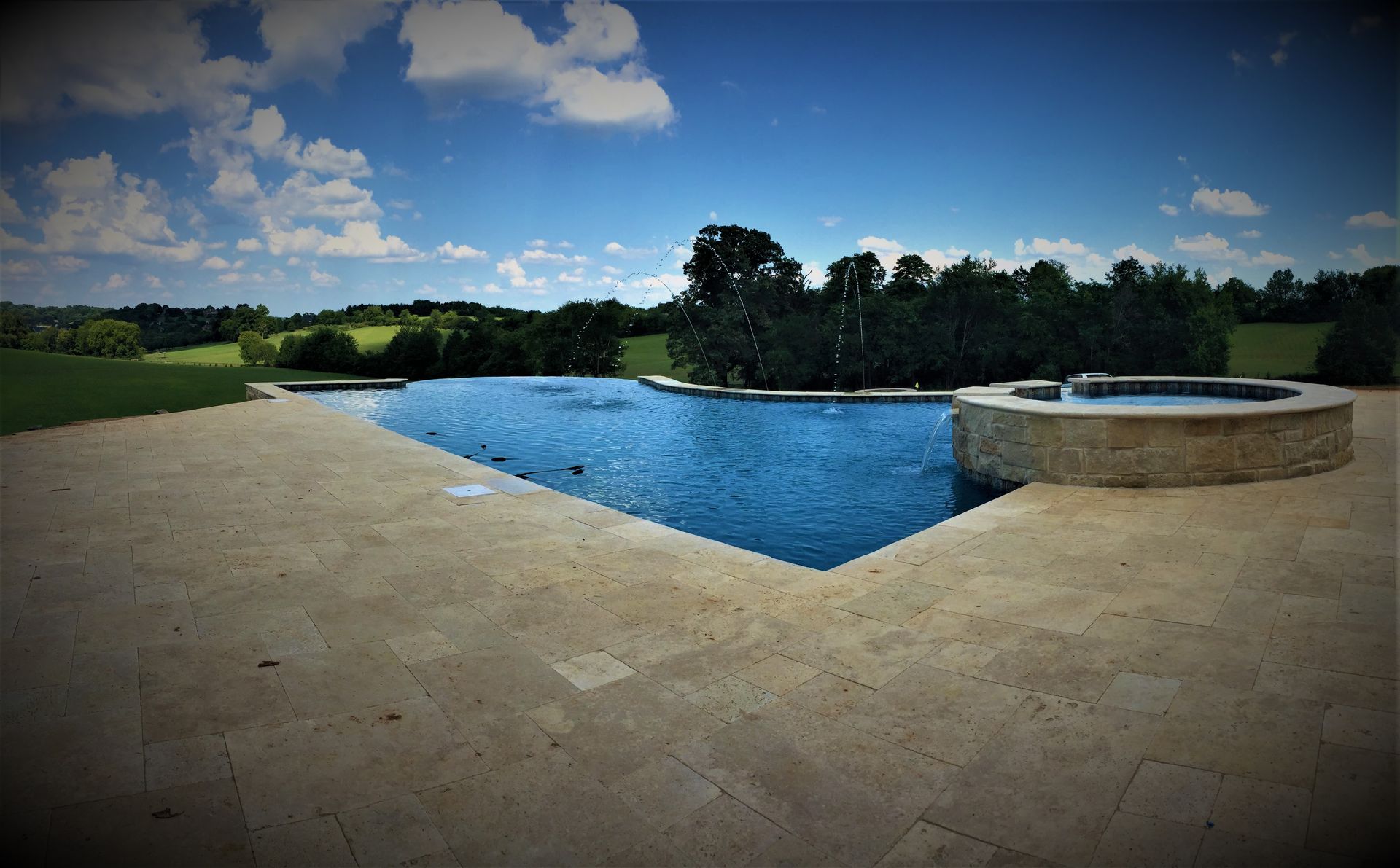 Blue swimming pool beside a circular hot tub on a stone patio, with trees and cloudy sky in the background