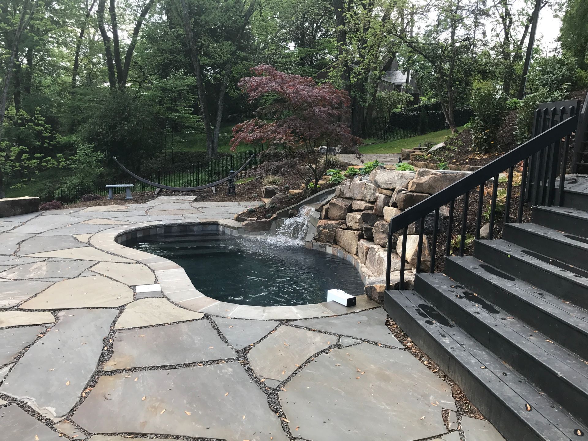 Backyard stone patio with a small pond, waterfall, and black staircase beside greenery