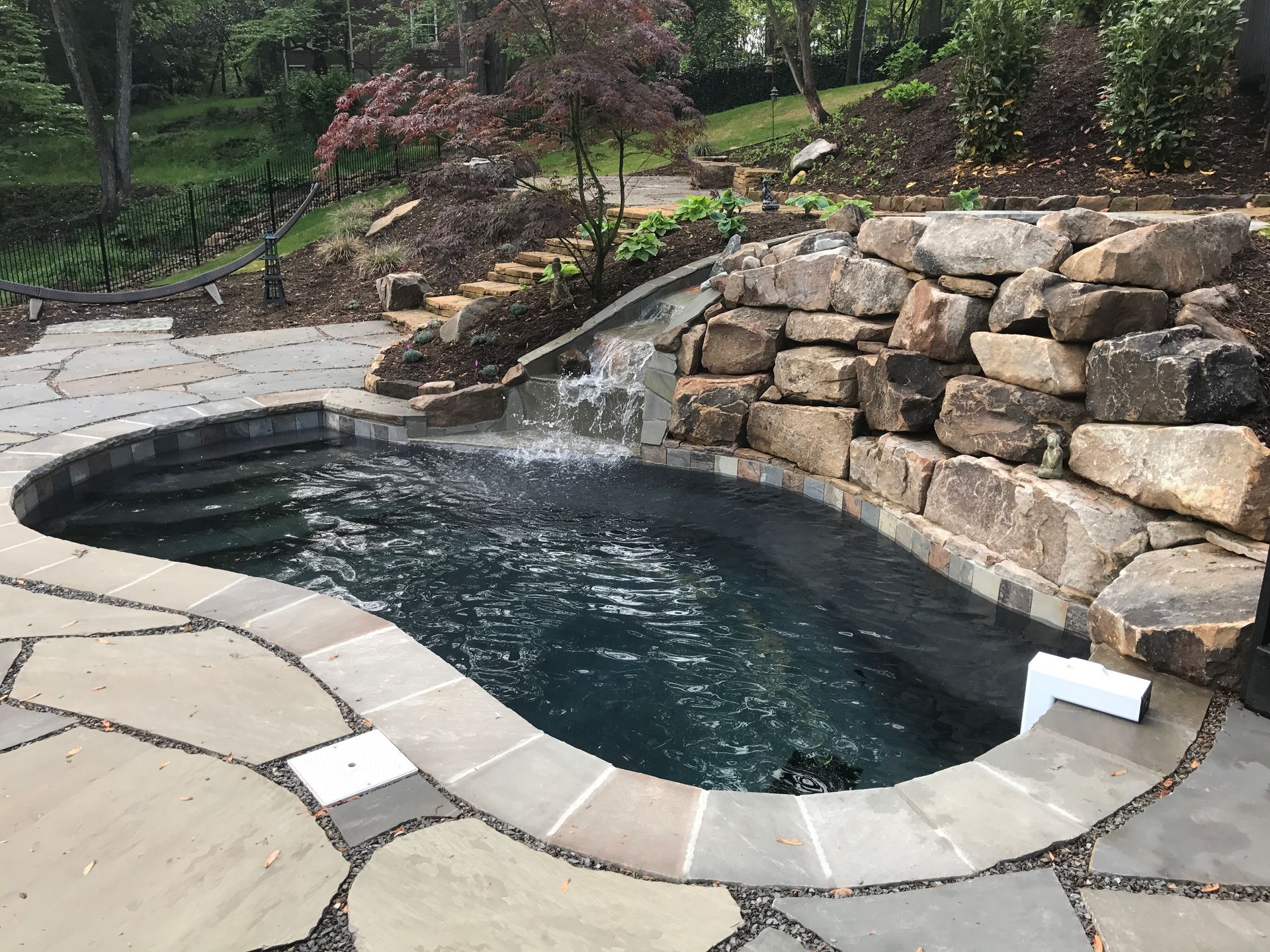 Backyard stone hot tub with dark water, surrounded by pavers and a stacked rock wall