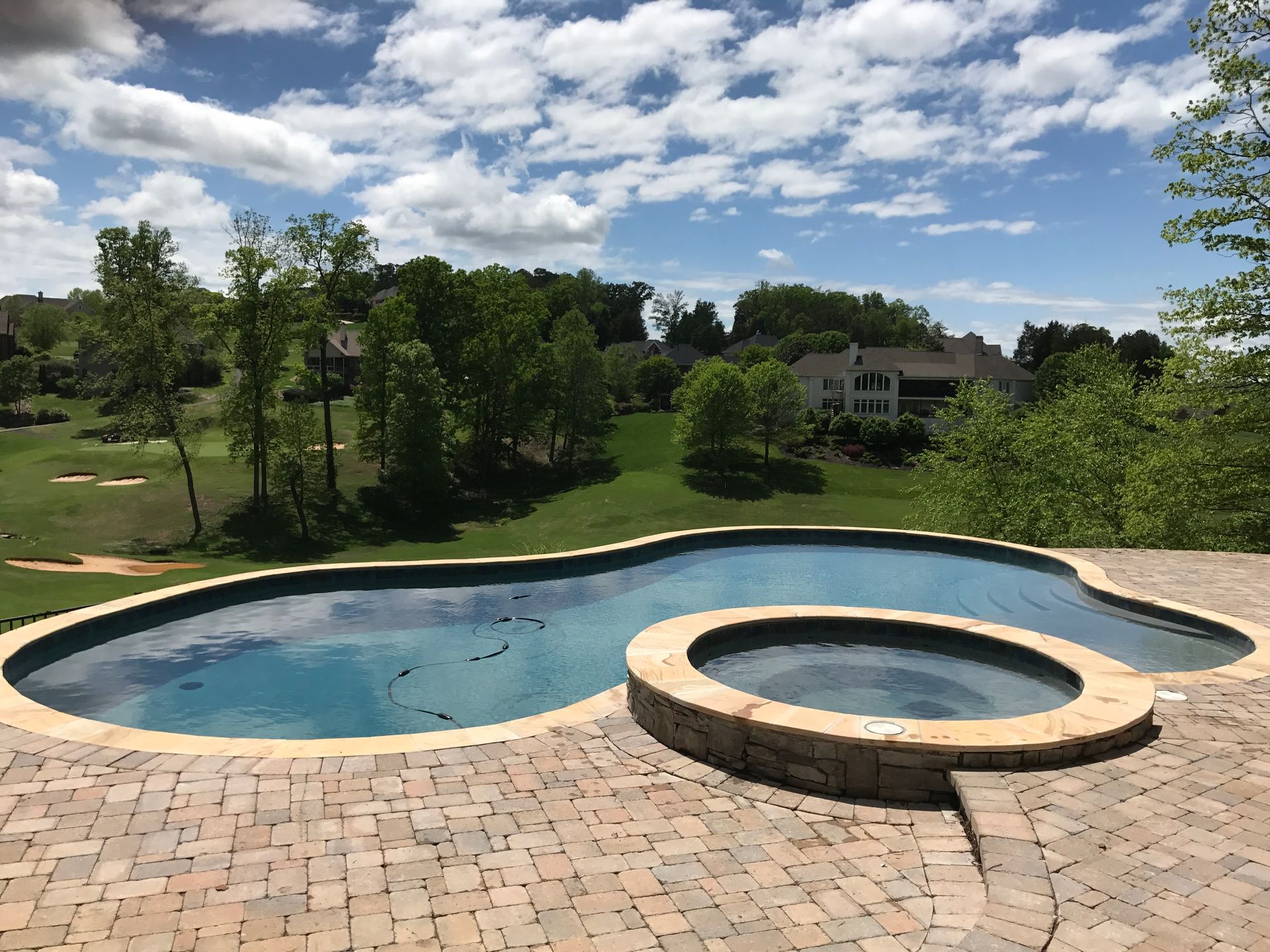 Backyard swimming pool and spa with brick patio, green lawn, trees, and a cloudy sky
