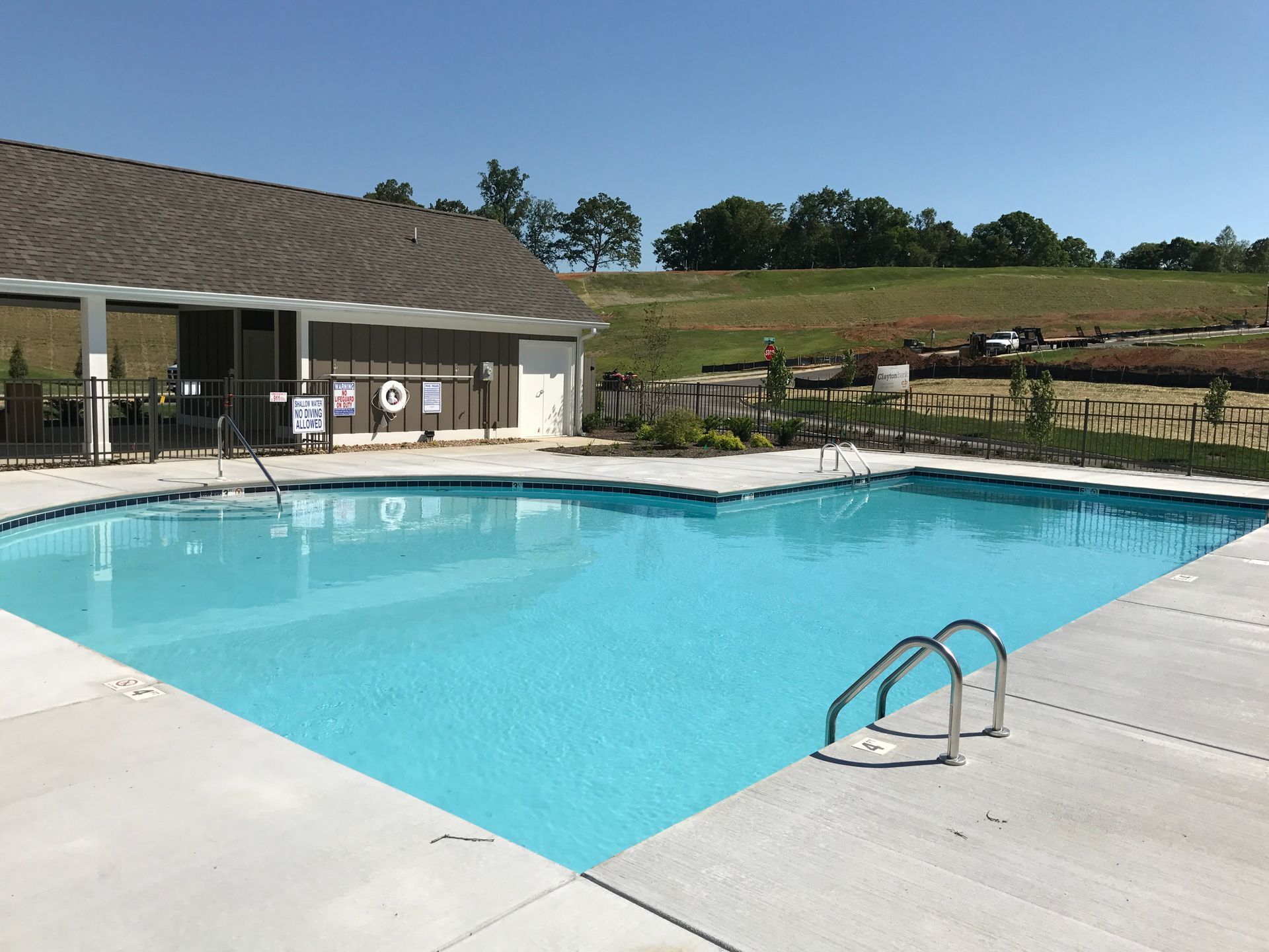 Outdoor swimming pool with clear blue water beside a clubhouse and fenced grassy hills.