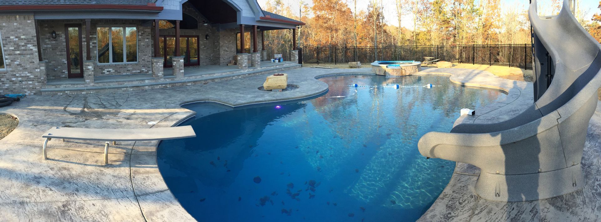 Outdoor stone pool with blue water beside a house, featuring a small waterfall and patio area.