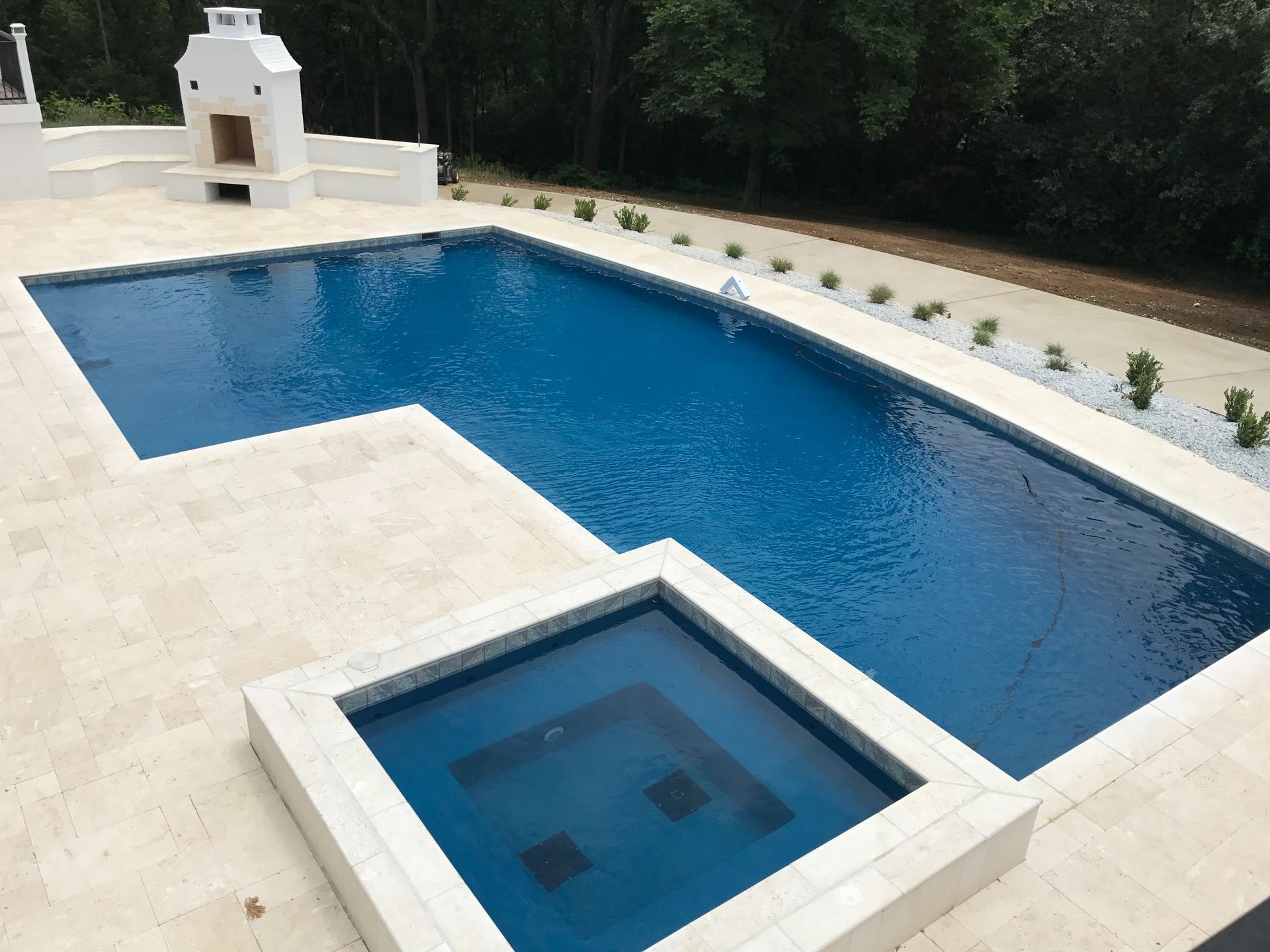 Modern outdoor pool with a small square hot tub, white stone deck, and greenery along the edge