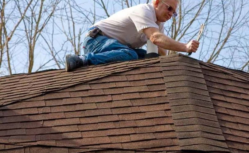 A man is kneeling on top of a roof holding a hammer.