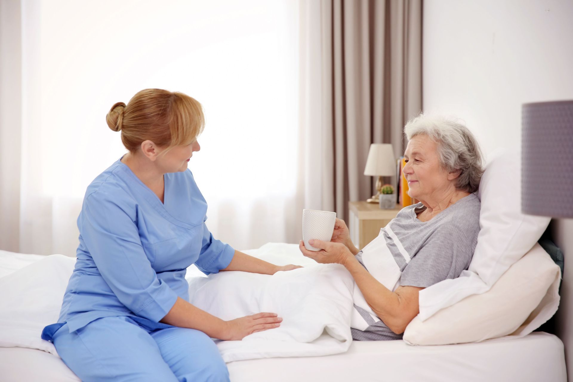 Nurse in blue scrubs offers a cup to an elderly patient resting in a hospital bed