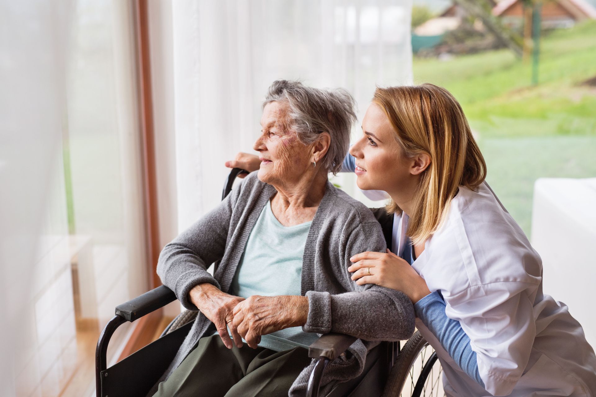 Older woman in a wheelchair smiling with a caregiver beside a bright window