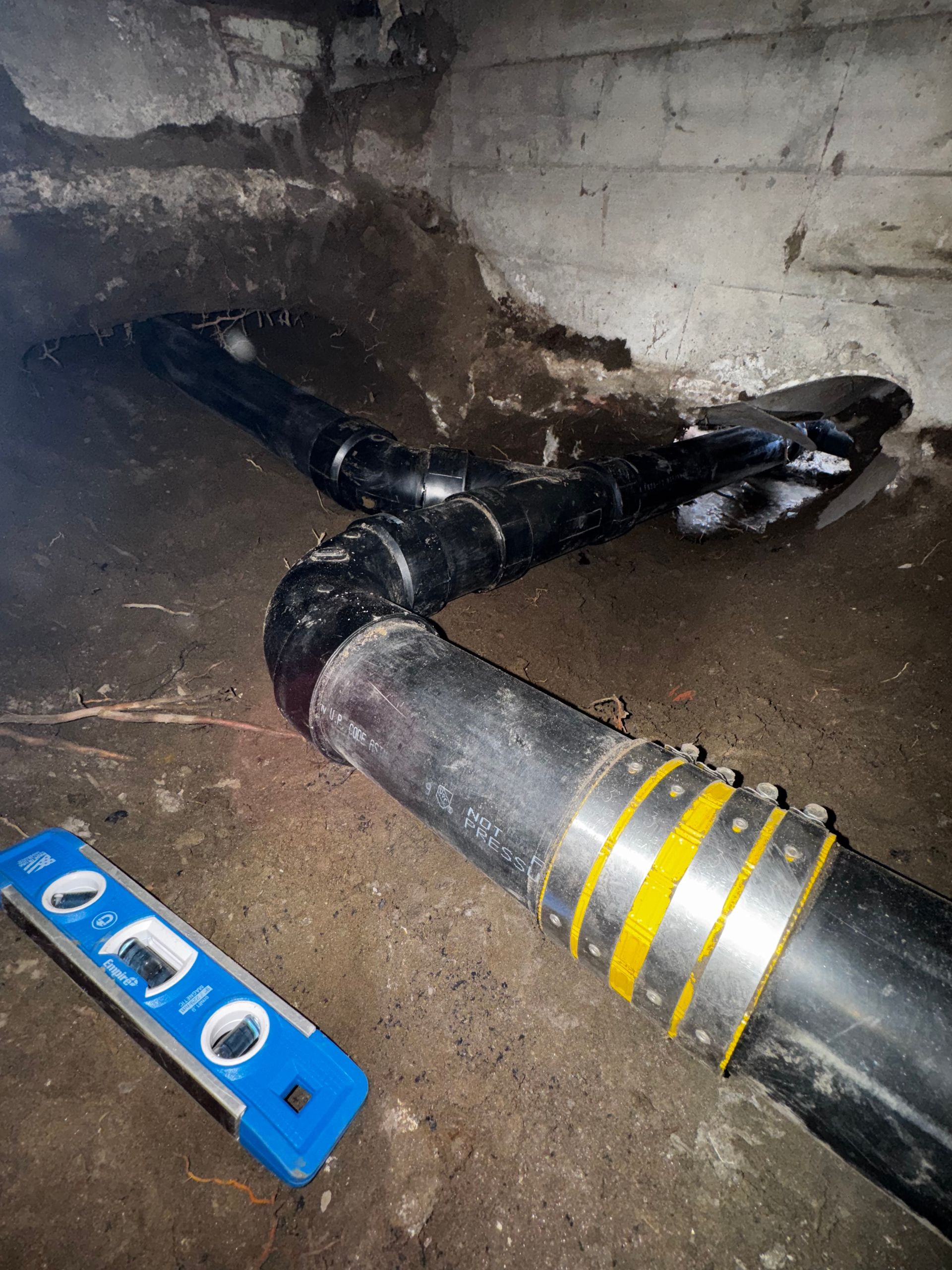 Black plumbing pipes with a yellow coupling in a concrete basement, next to a level tool.