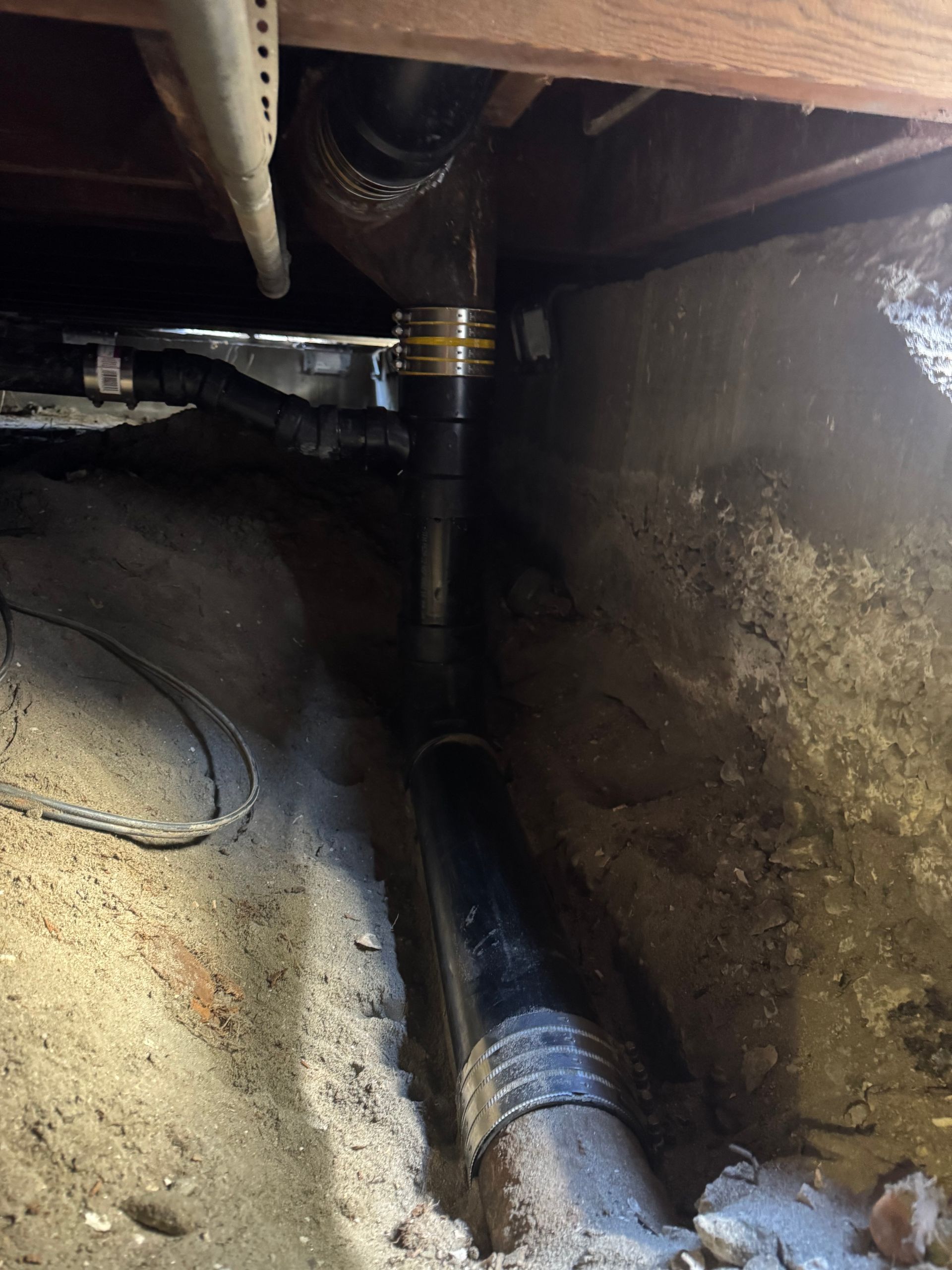 Black plumbing pipes in a dirt trench under a wooden structure. A black pipe connects to a Y-shaped pipe above.