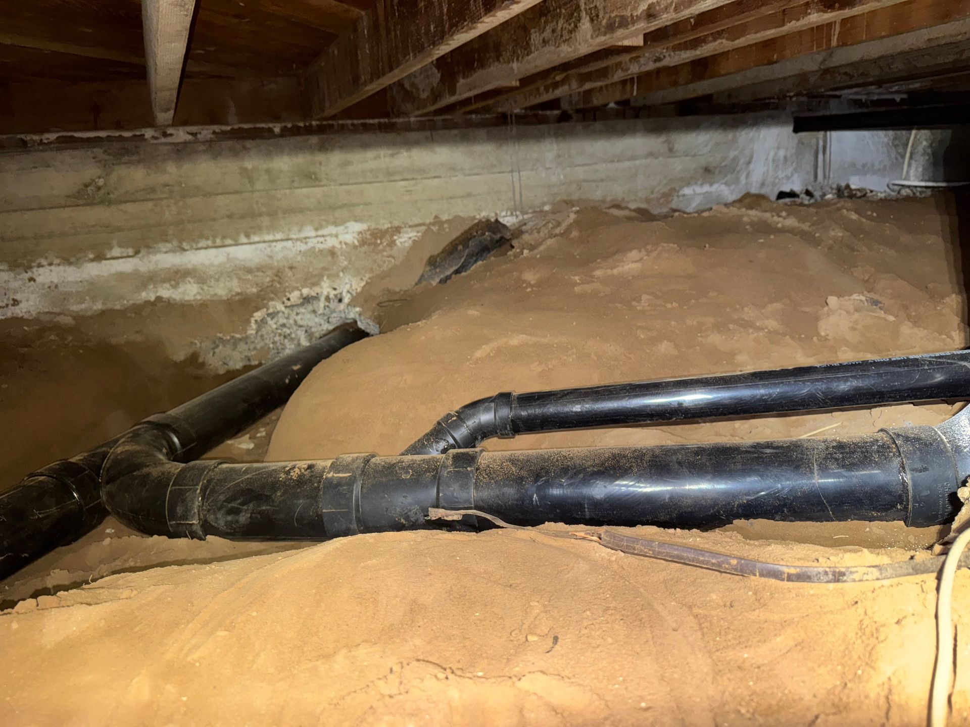 Black pipes run over dirt in a crawl space under a building. Wooden beams are above.