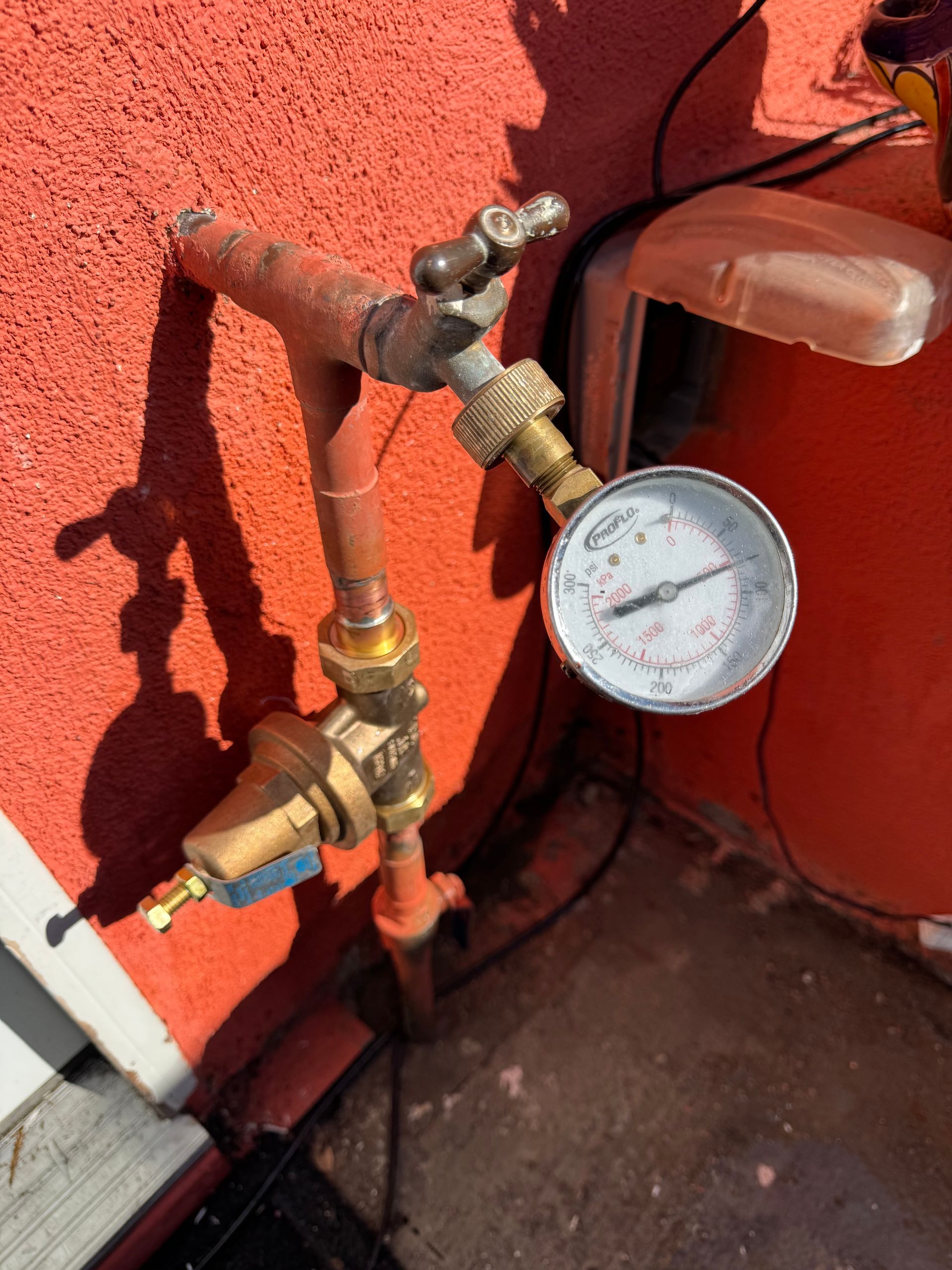 Water pressure gauge and pipes mounted on a red wall, outdoor setting.