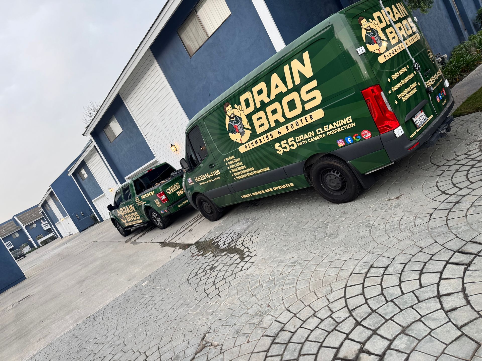 Green and gold Drain Bros service van and truck parked in front of blue apartment buildings.